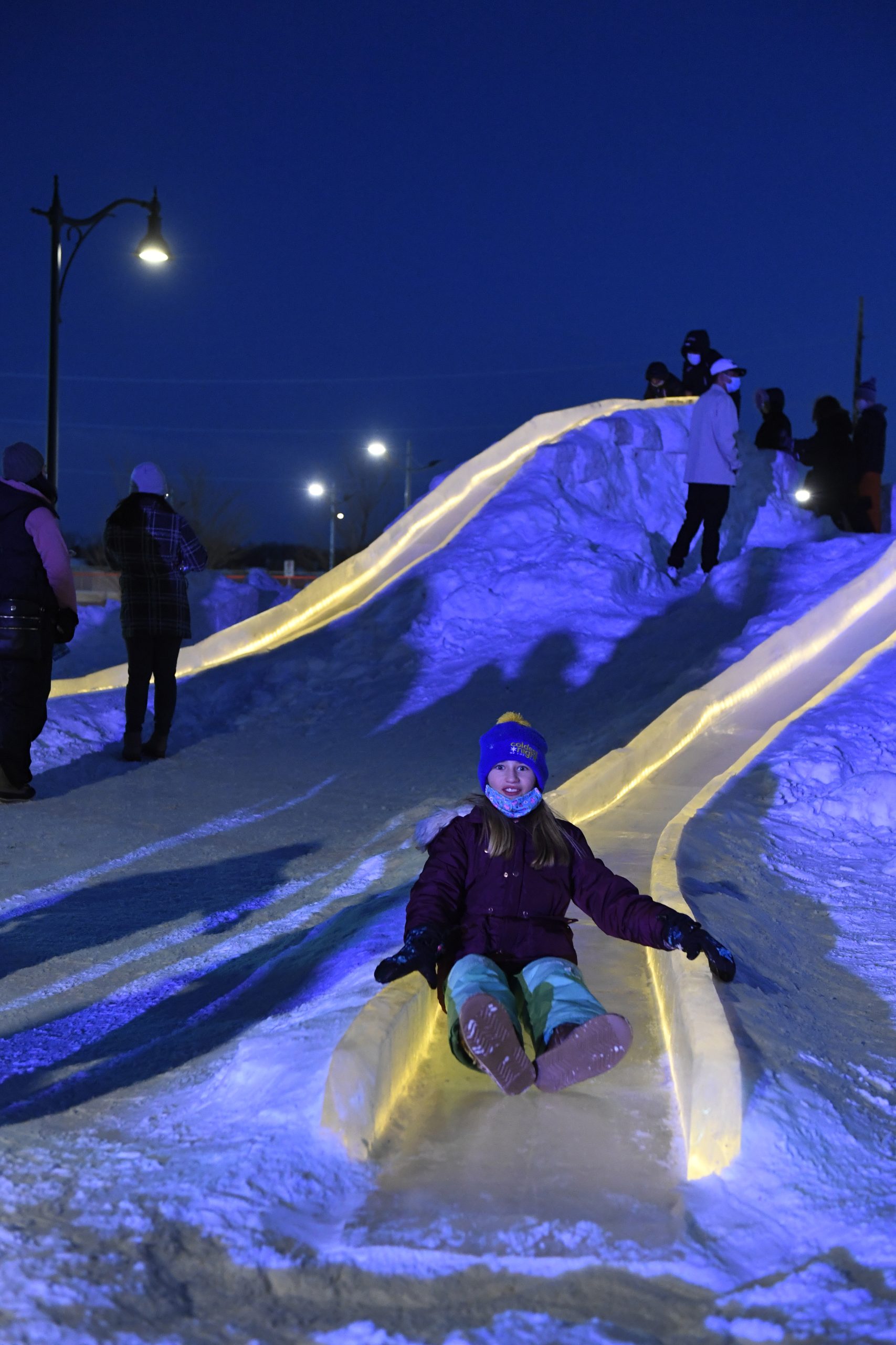 Child sliding down illuminated ice slide at night.